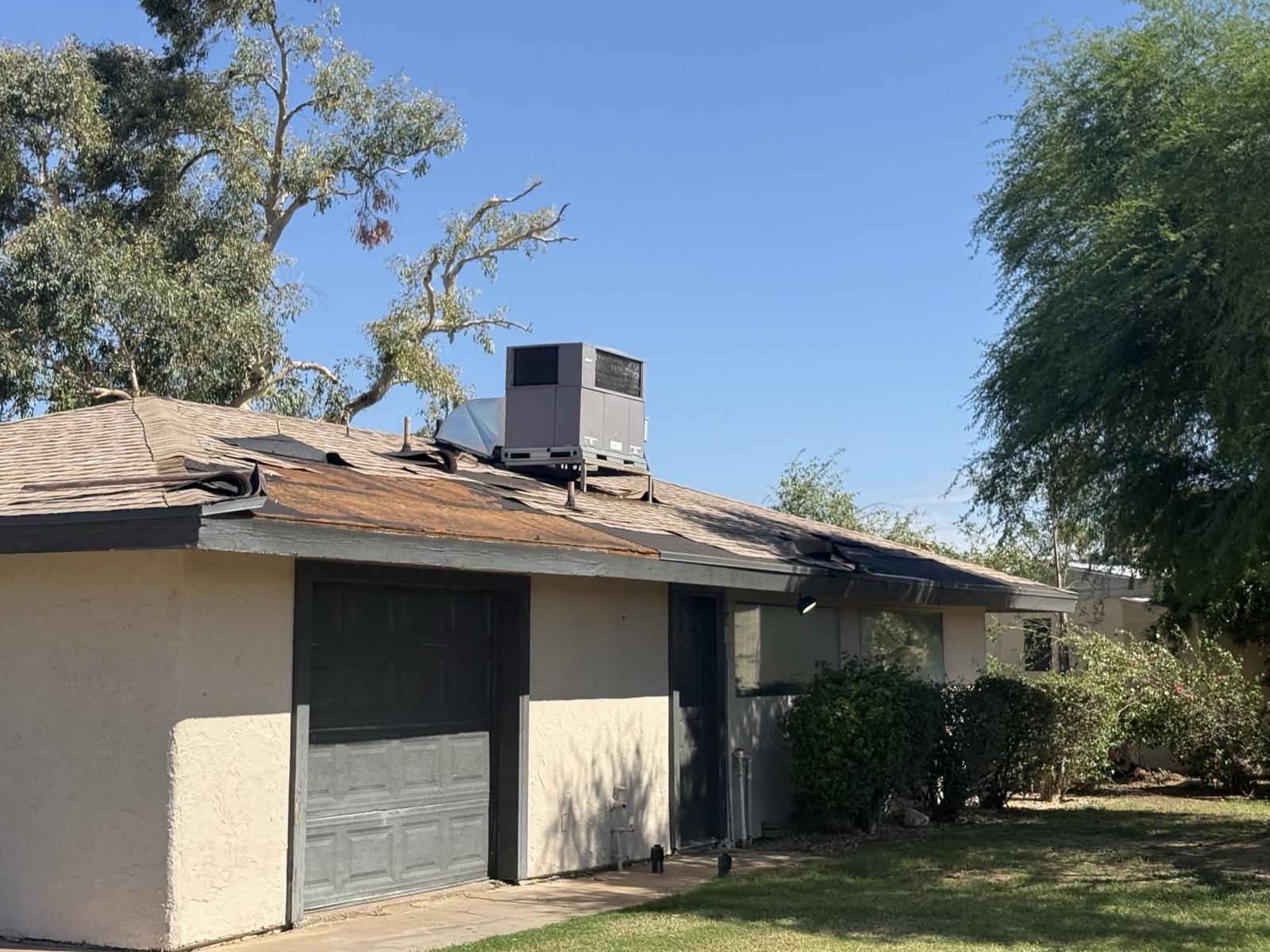 Before photo of a single-story commercial shop building in Tempe with a failing shingle roof, sections of shingles lifted and rolled back, and a packaged rooftop air conditioning unit
