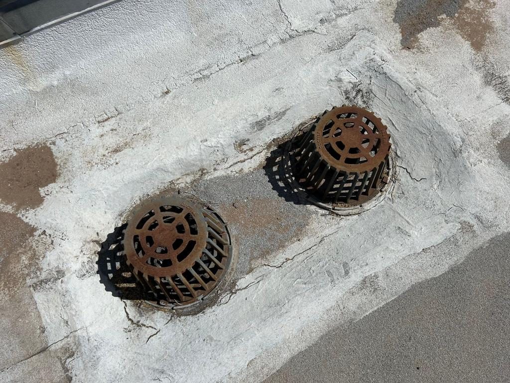 CS18 6 Before photo of two rusted cast iron roof drain strainers on a Phoenix cap sheet roof, set in a cracked sump patched over with layers of old mastic