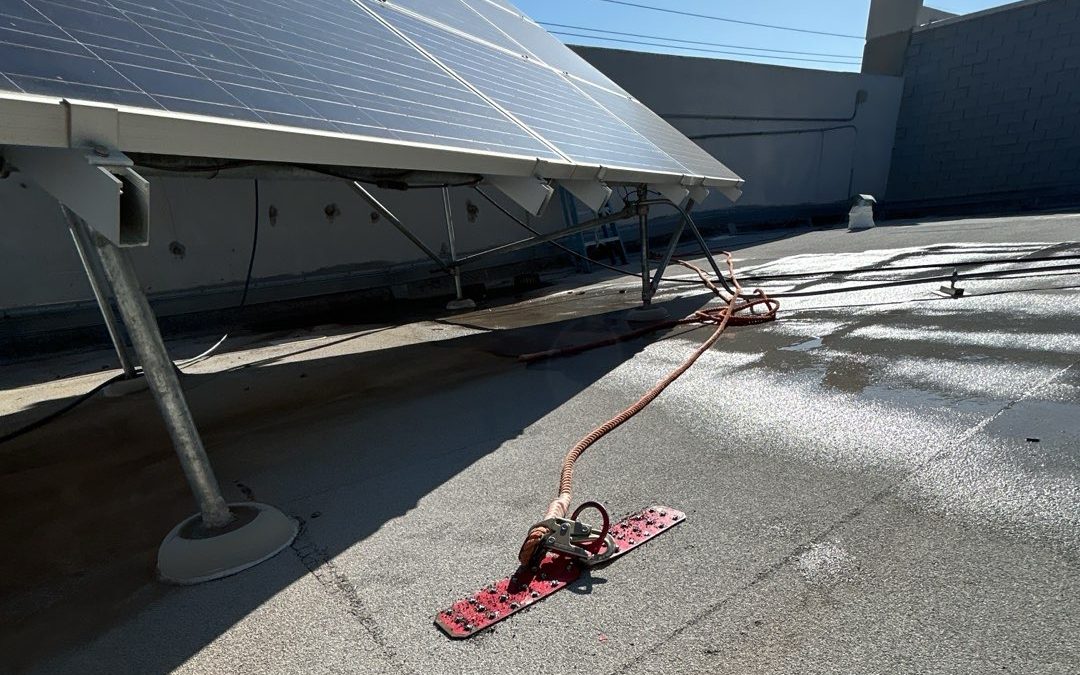 CS18 5 Progress photo of a pressure washer wand and hose lying on a granulated cap sheet roof beneath a rooftop solar array, with washed and unwashed areas side by side