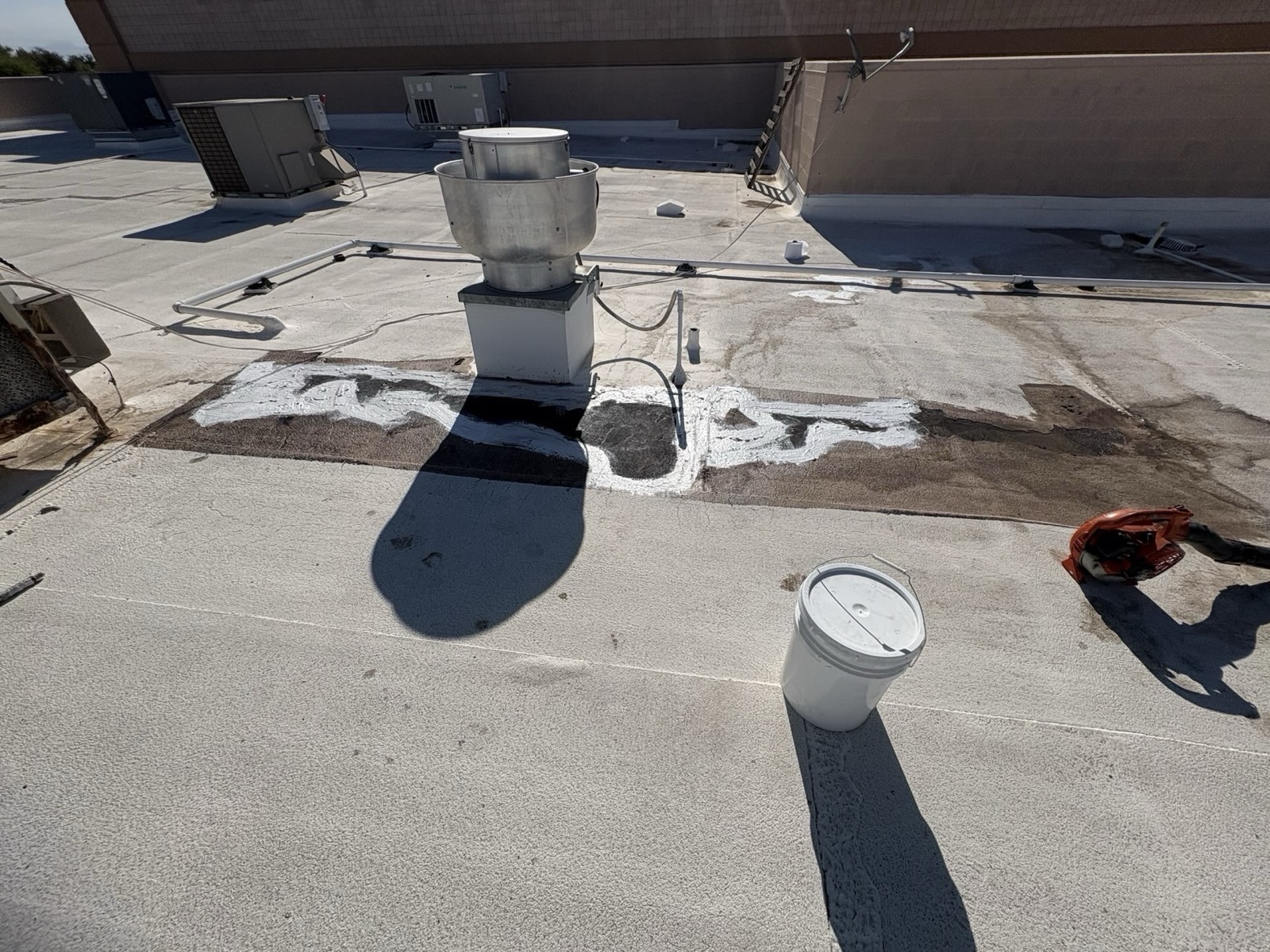 CS19 5 Maintenance work underway on a Gilbert commercial roof, with white sealant applied in bands around the curb of a rooftop exhaust unit over a dark runoff-stained area, beside a sealant pail and blower